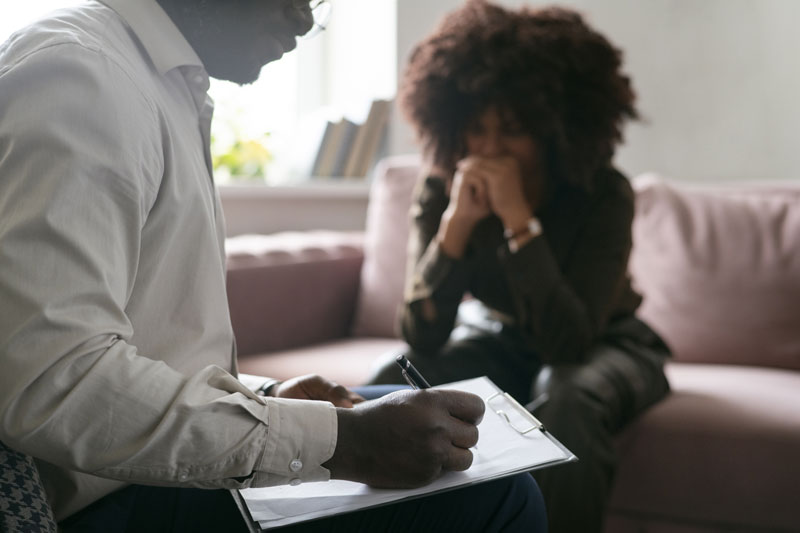 Therapist notes on a clipboard as a distressed client sits on a sofa seeking help in a living room
