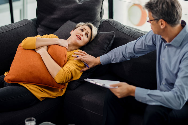 Woman on a couch grips an orange pillow distressed as a clinician reaches to help her with a clipboard nearby