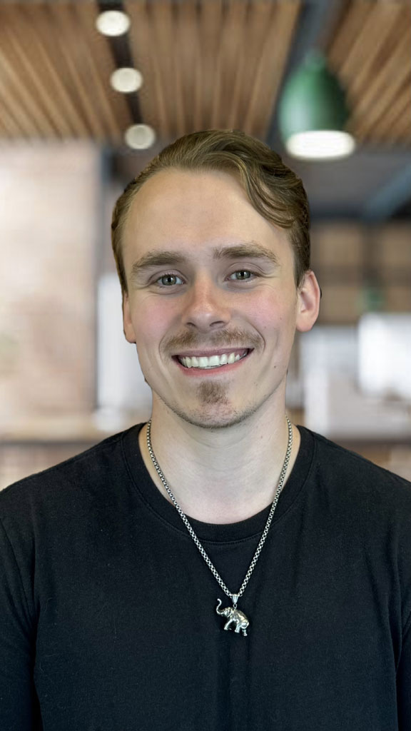 Smiling young man with short light brown hair wearing a black Tshirt and an elephant pendant necklace in a bright indoor setting