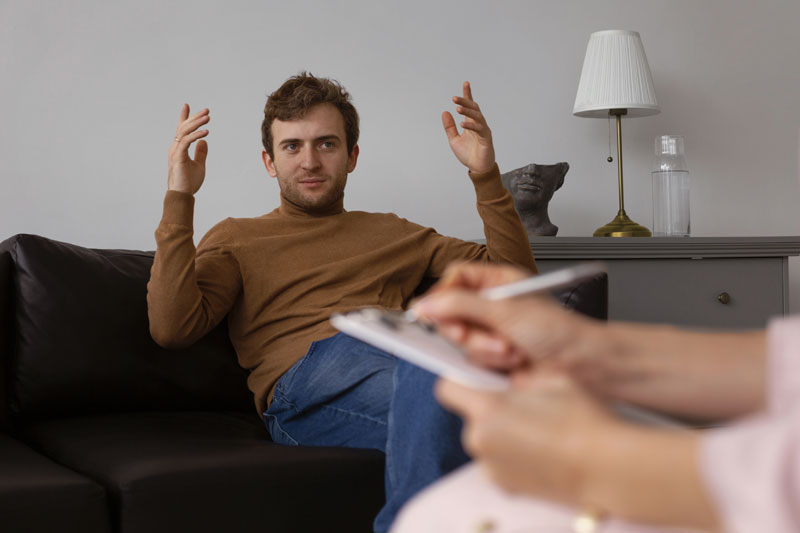 Man on a couch in a counseling session gesturing with his hands while the therapist takes notes in the foreground