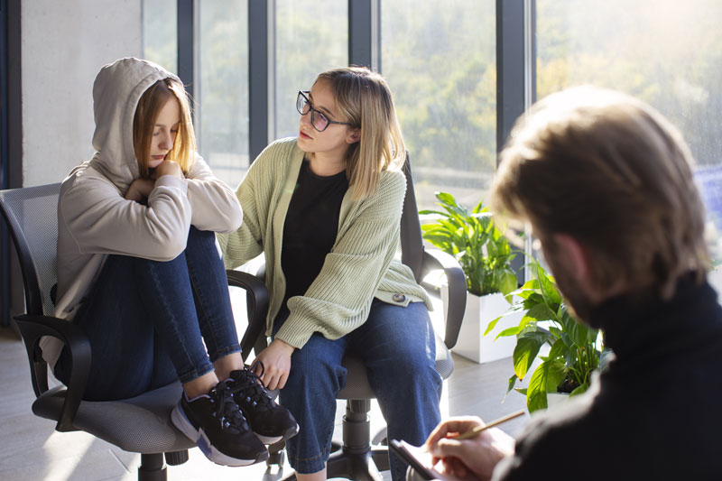 Three people in a bright office a young woman in a hoodie sits with knees drawn up while another woman with glasses talks to her a man in the foreground writes in a notebook