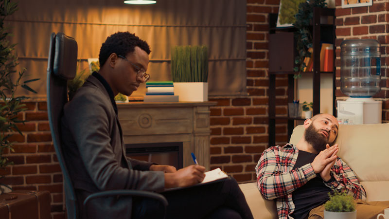 Therapist sits in a chair taking notes while a bearded client reclines on a sofa during a counseling session