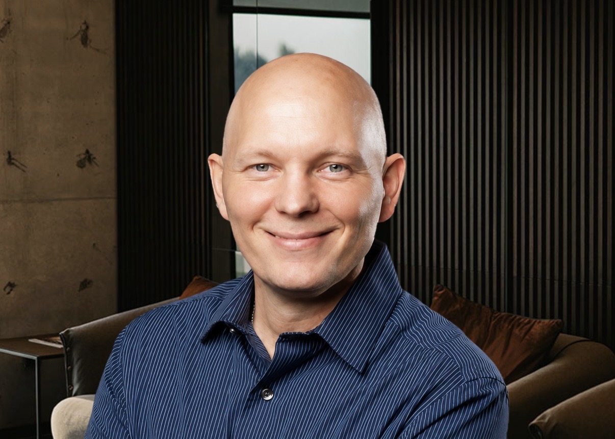 Smiling bald man in a blue striped shirt seated in a modern lounge with dark vertical panels and brown cushions
