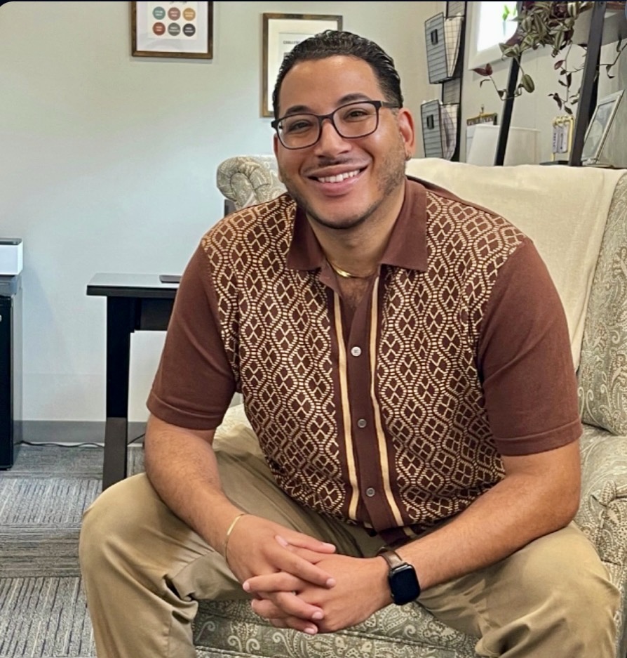 Smiling man wearing glasses sits on a patterned couch in an office wearing a brown geometric pattern shirt and tan pants